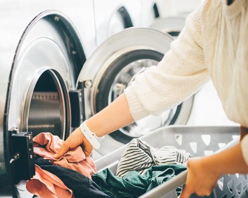 Interior of small laundromat in daylight. Close-up female holding basket. Girl loading dirty clothes inside drum and closing door. Self-service concept